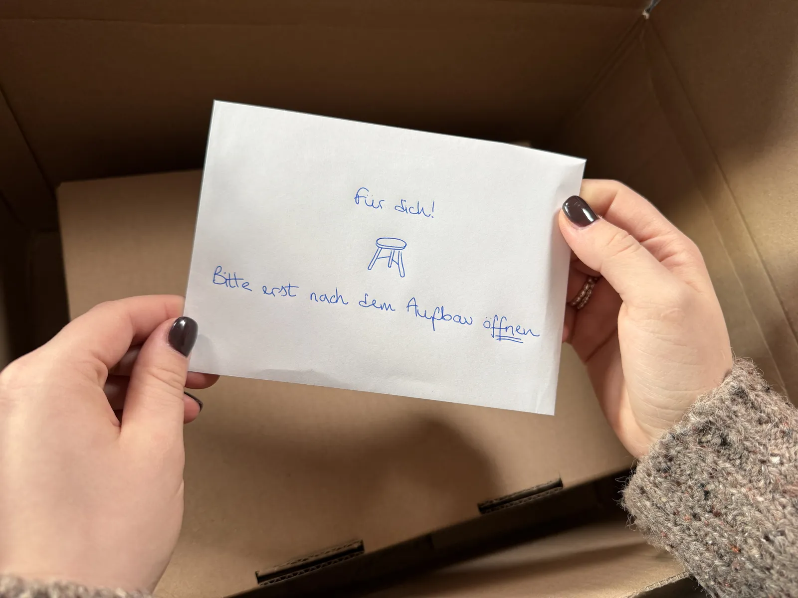 Hände halten einen handbeschrifteten Umschlag „Für dich! Bitte erst nach dem Aufbau öffnen“ über dem offenen Versandkarton