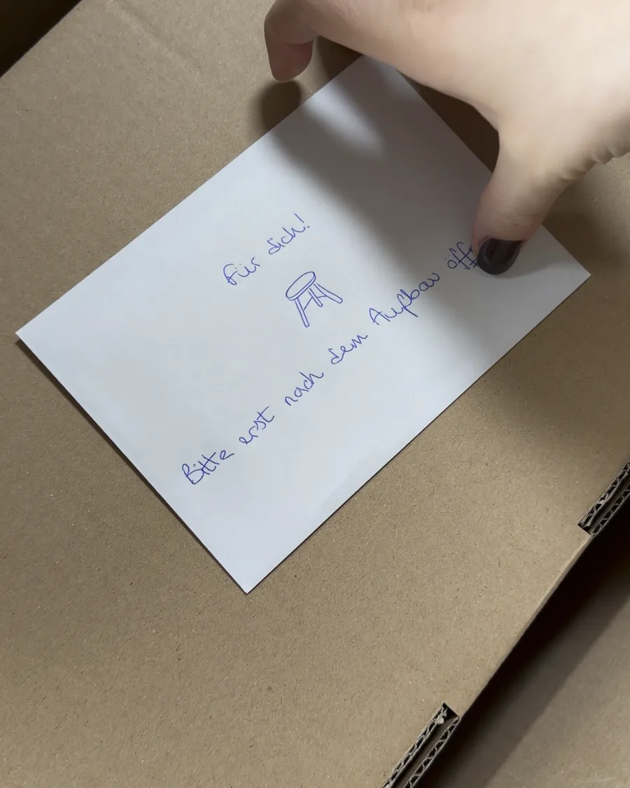 Offener Versandkarton, oben liegt ein Umschlag: Für dich! Bitte erst nach dem Aufbau öffnen