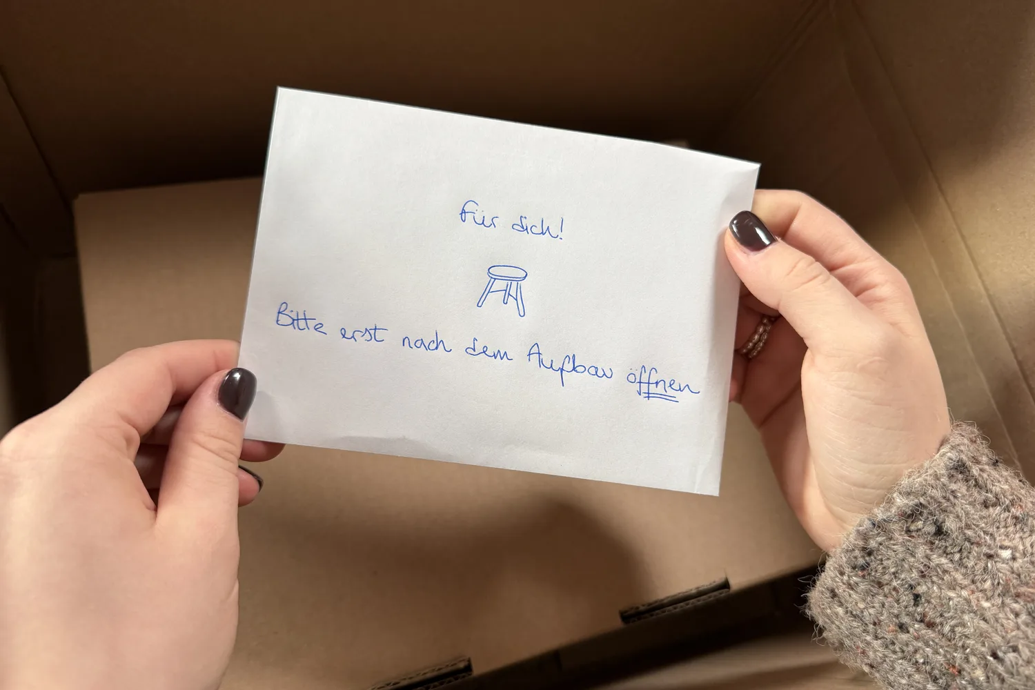 Hände halten über einem offenen Versandkarton einen handbeschrifteten Umschlag: Für dich! Bitte erst nach dem Aufbau öffnen