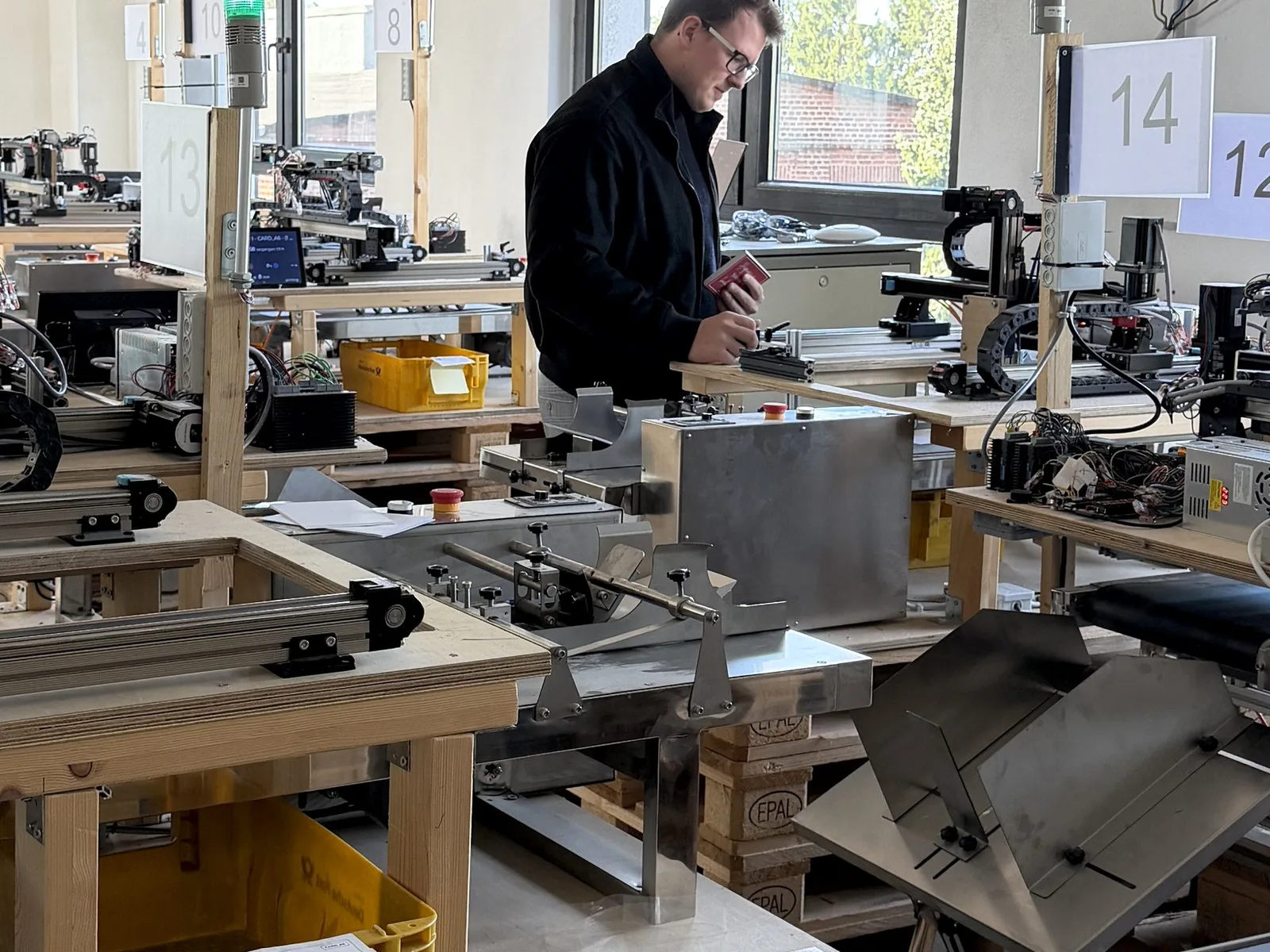 Werkstatt mit mehreren Schreibrobotern auf Holzgestellen, ein Mitarbeiter prüft eine Maschine